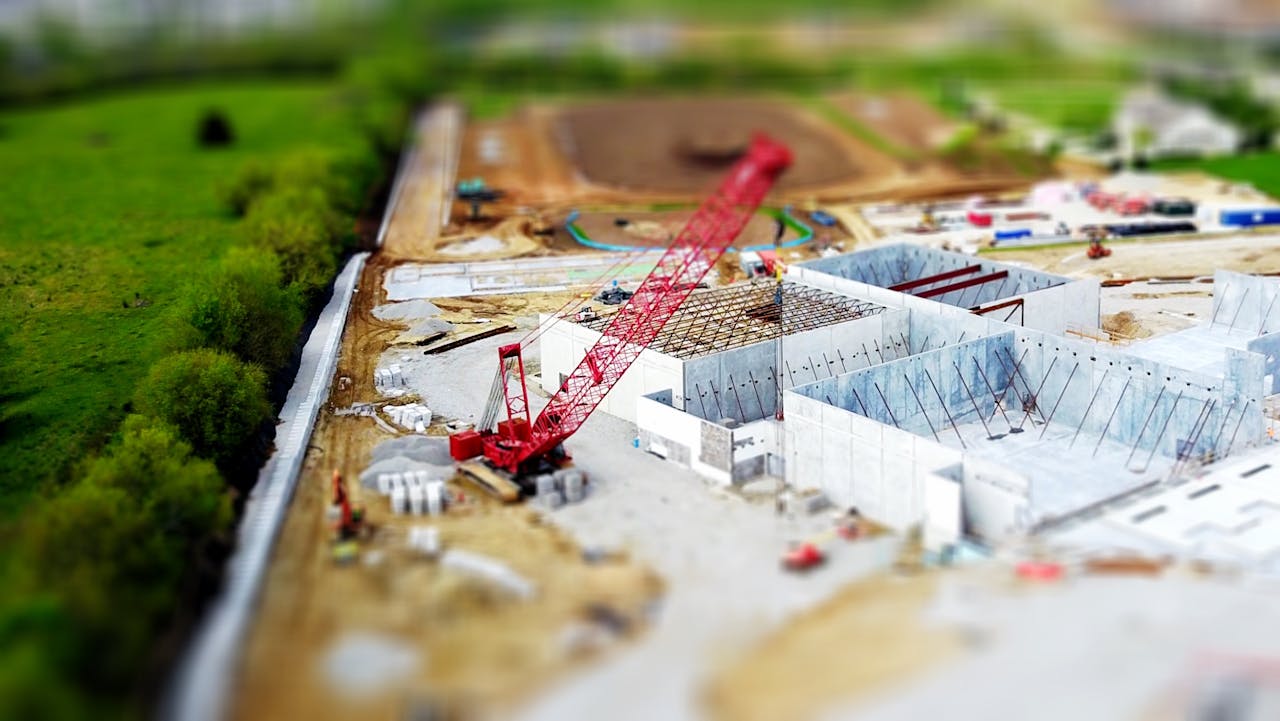 our-story-02 Aerial view of a construction site with a red crane amidst green fields and concrete buildings.